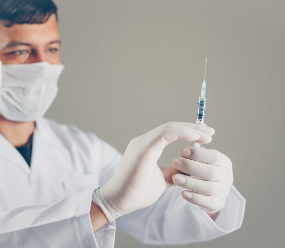 Doctor with gloves and mask looking at syringe and holding syringe with his hands on a gray background. side view.